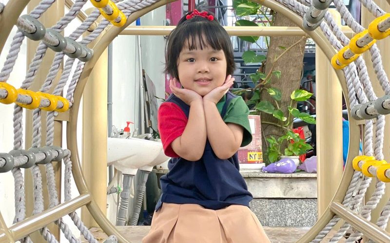 Bé gái mặc đồng phục chống tay lên má cười trong khu leo trèo