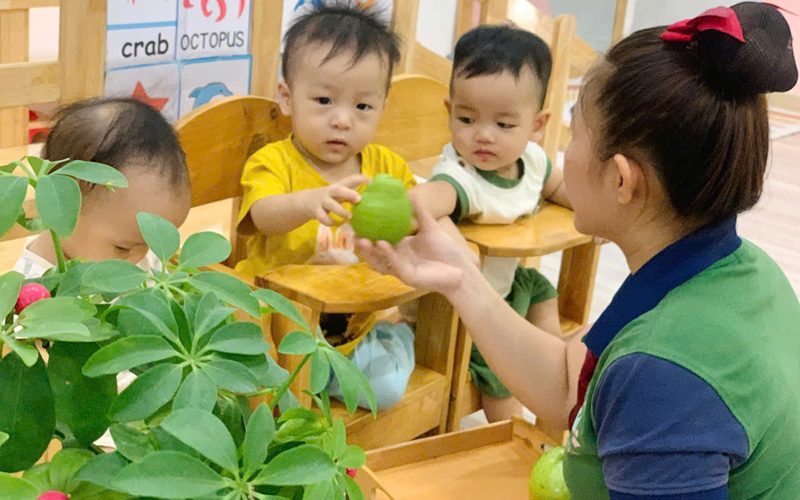Cô đưa quả cho hai bé nhỏ ngồi ghế ăn trong lớp nhà trẻ