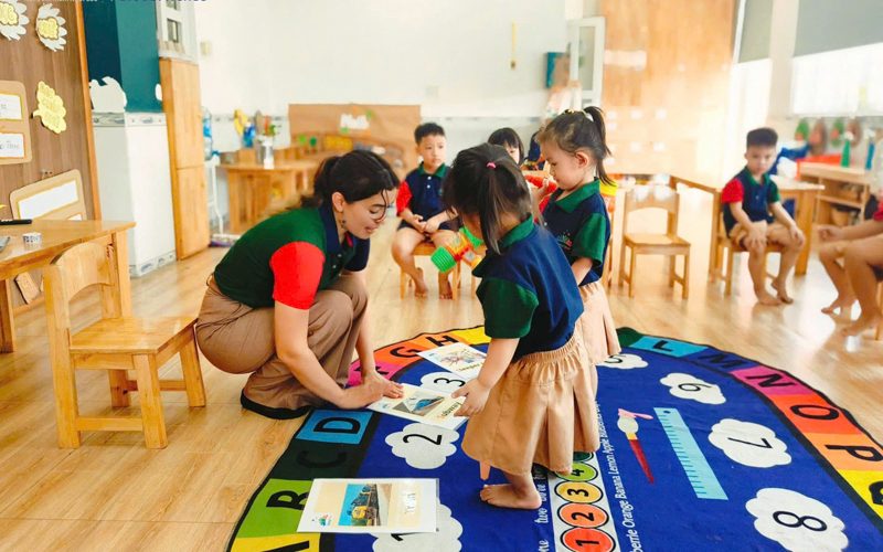 Cô ngồi xuống ngang tầm bé trên tấm thảm chữ cái và chữ số